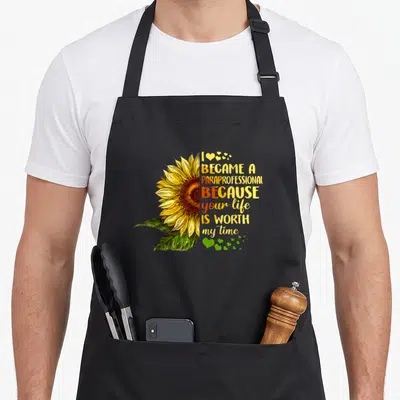 Became cute flamingo apron - Sunflower illustration with I became a paraprofessional text and scattered hearts — shown on a black cotton canvas apron against a white background