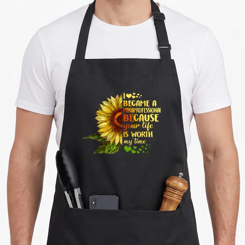 Became cute flamingo apron - Sunflower illustration with I became a paraprofessional text and scattered hearts — shown on a black cotton canvas apron against a white background