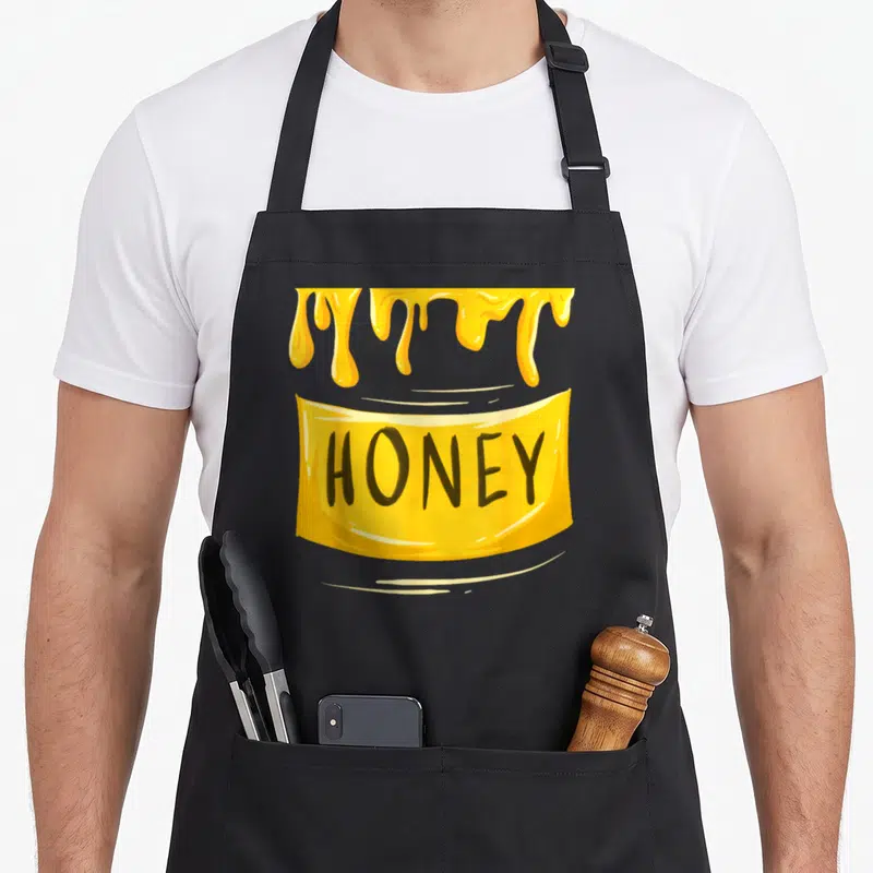 Sweet funny halloween aprons - Dripping honey above yellow banner with black HONEY text in cartoon style — shown on a black cotton canvas apron against a white background