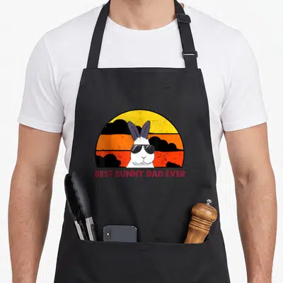 Bunny rabbit daddy apron - White bunny wearing sunglasses in front of a yellow orange and black retro sun with red text — shown on a black cotton canvas apron against a white background