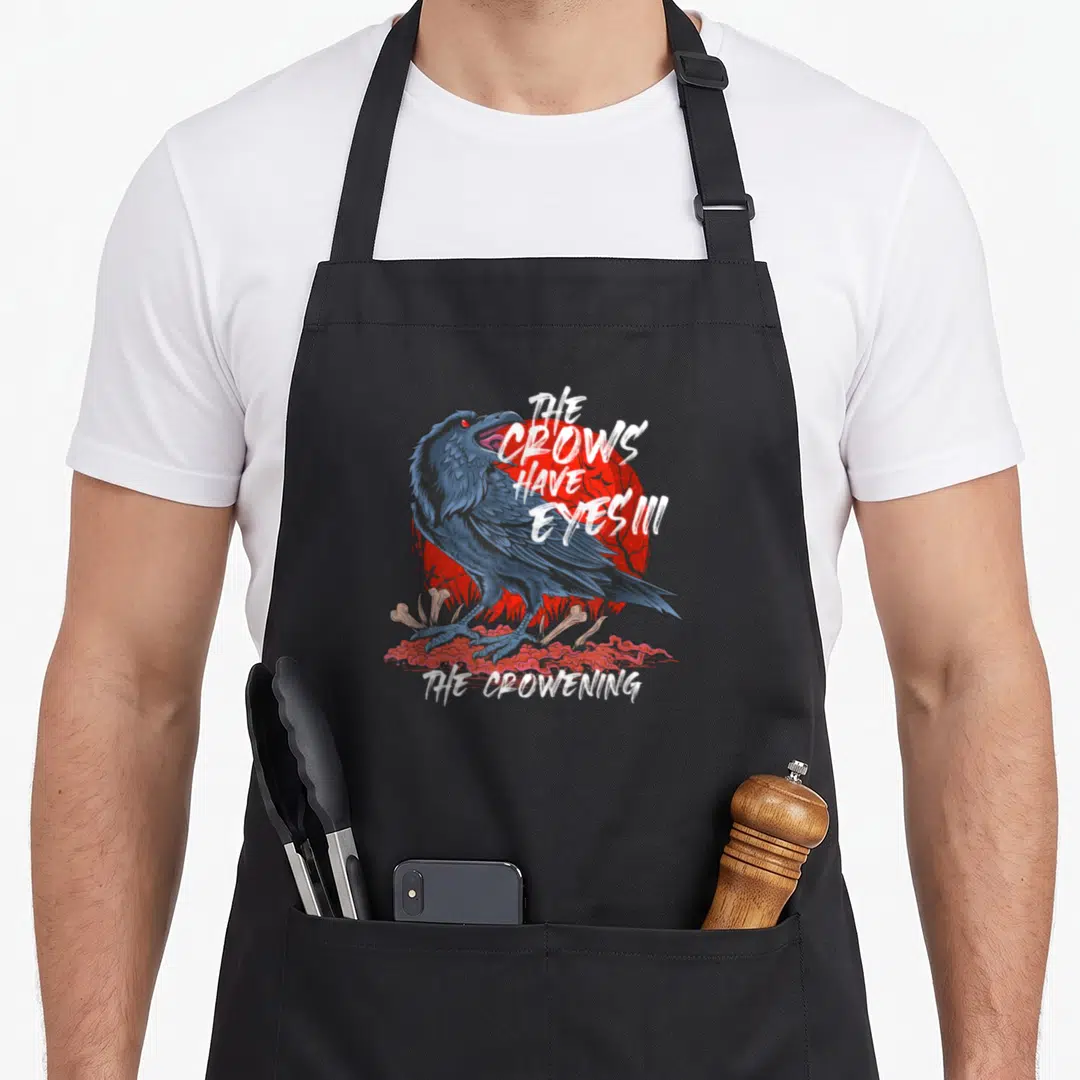 Eye crows before apron - Dark blue crow on red sun with bones and white text reading The Crows Have Eyes III — shown on a black cotton canvas apron against a white background