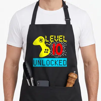 10th birthday gamer video game apron - Pixel yellow controller beside red number 10 with white rays and blue UNLOCKED — shown on a black cotton canvas apron against a white background