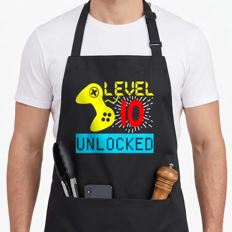 10th birthday gamer video game apron - Pixel yellow controller beside red number 10 with white rays and blue UNLOCKED — shown on a black cotton canvas apron against a white background
