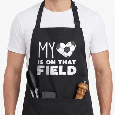 Field soccer football apron - Distressed white text MY IS ON THAT FIELD with soccer ball heart on grey background — shown on a black cotton canvas apron against a white background