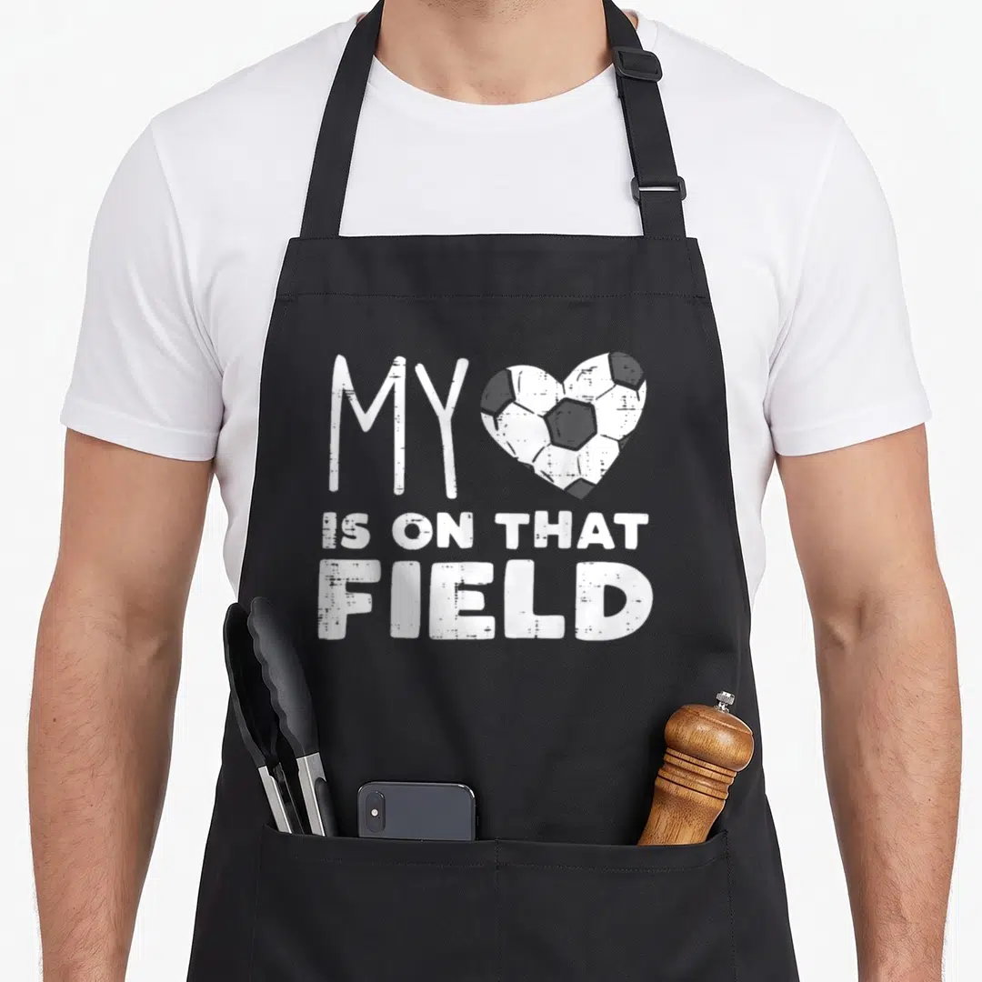 Field soccer football apron - Distressed white text MY IS ON THAT FIELD with soccer ball heart on grey background — shown on a black cotton canvas apron against a white background