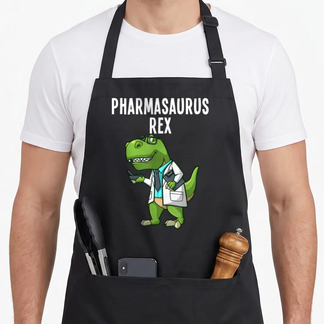 Dinosaur pharmacist apron - Green cartoon T-Rex in lab coat holding mortar and clipboard under stacked white text — shown on a black cotton canvas apron against a white background