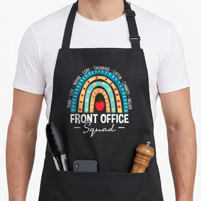 Squad school office apron - Rainbow with red apples and words TEACH LOVE INSPIRE LEAD ENCOURAGE LISTEN CONNECT INCLUDE — shown on a black cotton canvas apron against a white background