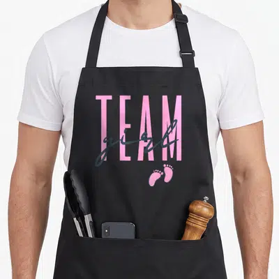 Baby shower apron or apron - Pink TEAM text with grey girl script and two pink footprints on grey background — shown on a black cotton canvas apron against a white background