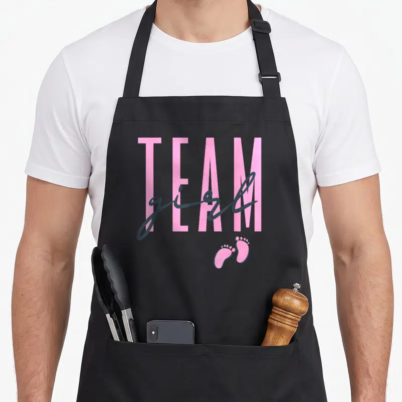Baby shower apron or apron - Pink TEAM text with grey girl script and two pink footprints on grey background — shown on a black cotton canvas apron against a white background