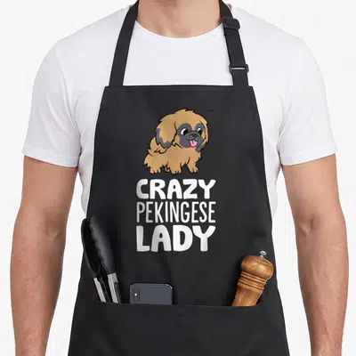 Pekingese puppy dog apron - Cartoon tan Pekingese dog above white CRAZY PEKINGESE LADY text on black fabric — shown on a black cotton canvas apron against a white background