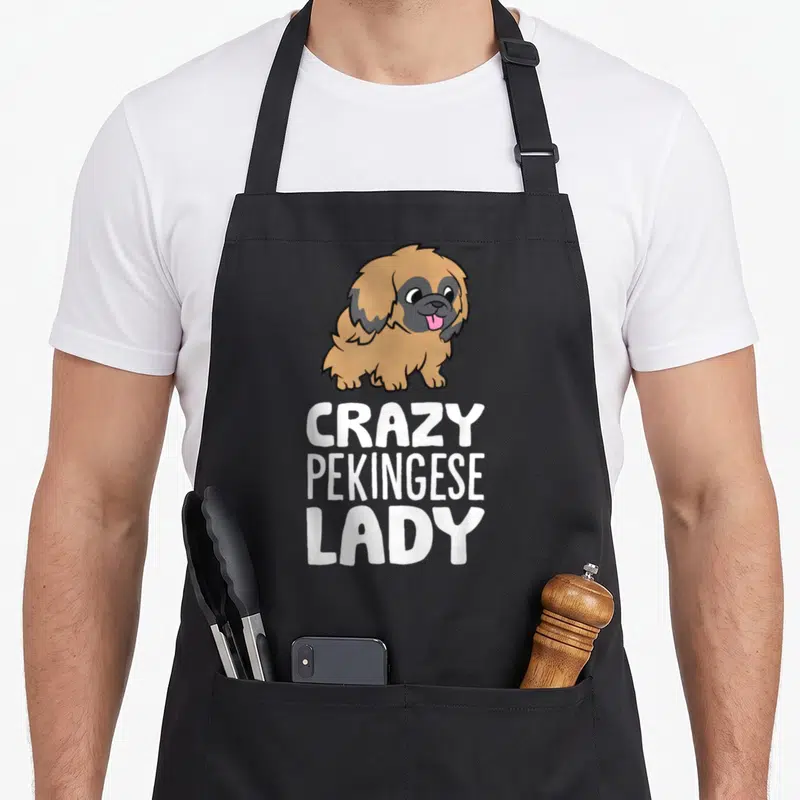 Pekingese puppy dog apron - Cartoon tan Pekingese dog above white CRAZY PEKINGESE LADY text on black fabric — shown on a black cotton canvas apron against a white background