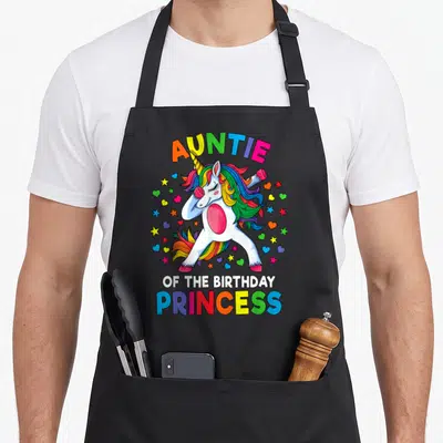 Auntie princess apron - Dabbing unicorn with rainbow hair and stars under arched text — shown on a black cotton canvas apron against a white background