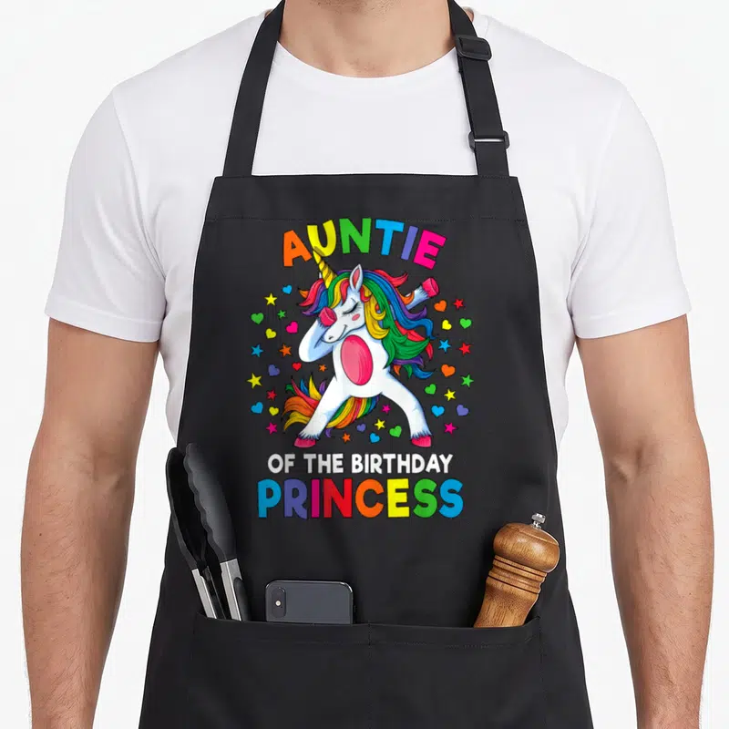 Auntie princess apron - Dabbing unicorn with rainbow hair and stars under arched text — shown on a black cotton canvas apron against a white background