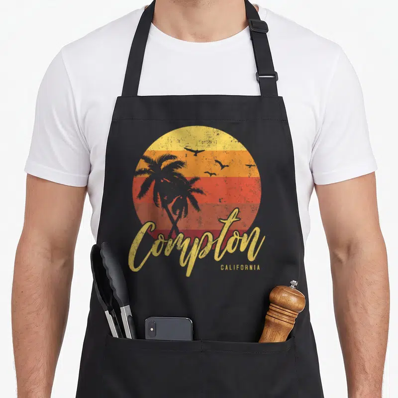 Cool california compton apron - Retro sunset with palm tree silhouettes and script Compton California text on black bib — shown on a black cotton canvas apron against a white background
