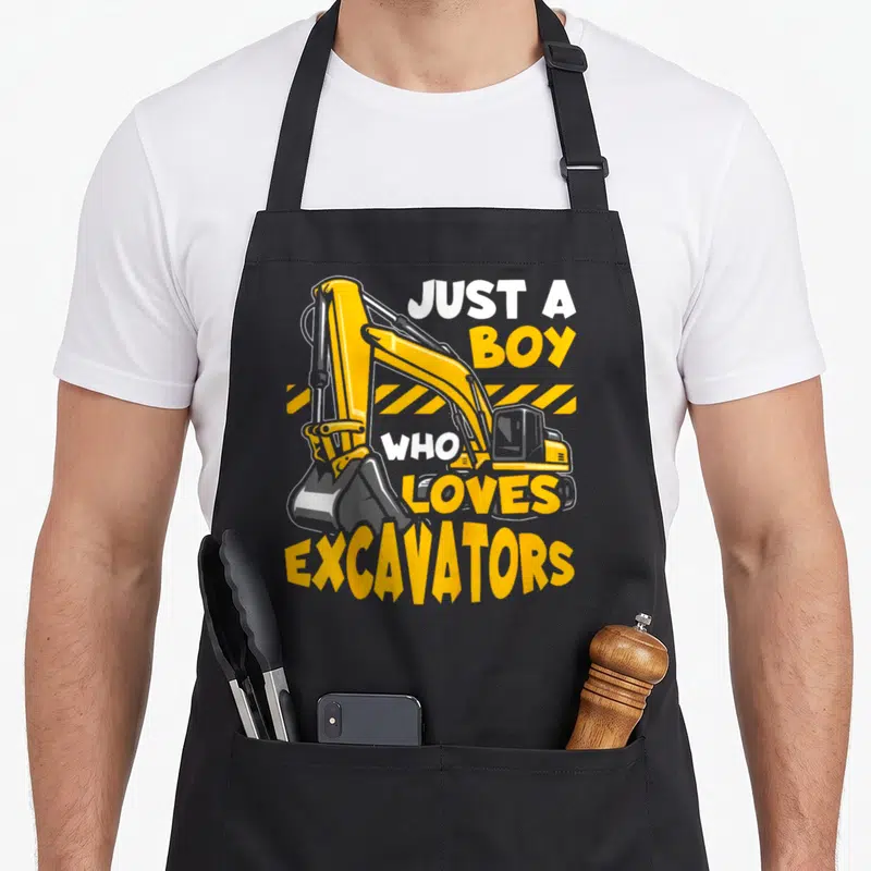 Construction vehicle apron - Yellow cartoon excavator with hazard stripes and text JUST A BOY WHO LOVES EXCAVATORS — shown on a black cotton canvas apron against a white background