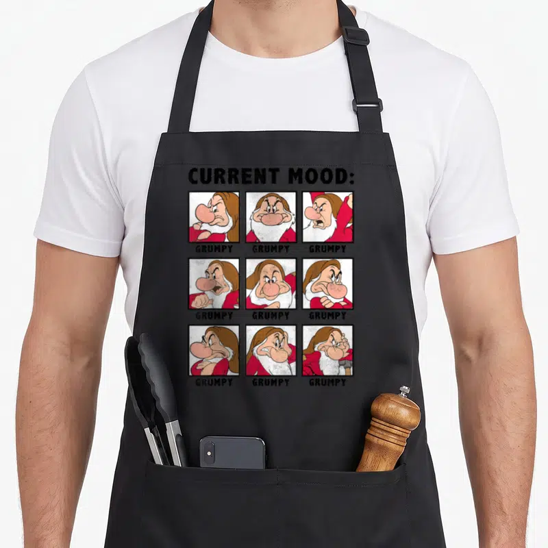 Current snow white apron - Three by three grid of grumpy dwarf cartoon faces with current mood label and grumpy captions — shown on a black cotton canvas apron against a white background