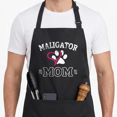 Funny maligator belgian malinois apron - Arched MALIGATOR text above pink heart overlapping white paw print and MOM — shown on a black cotton canvas apron against a white background