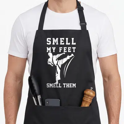 Smell taekwondo apron - White martial artist silhouette kicking between text SMELL MY FEET SMELL THEM on black — shown on a black cotton canvas apron against a white background