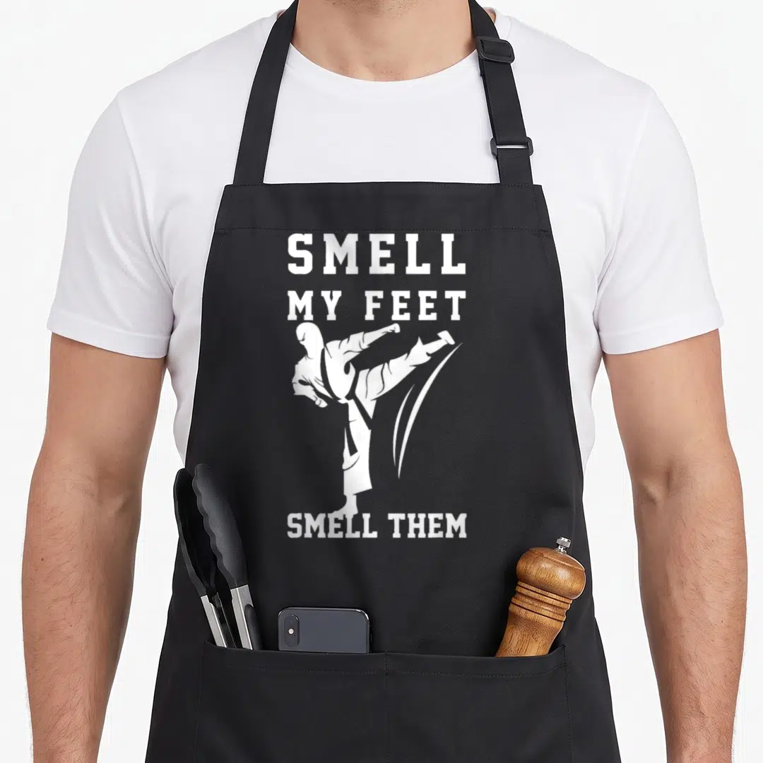Smell taekwondo apron - White martial artist silhouette kicking between text SMELL MY FEET SMELL THEM on black — shown on a black cotton canvas apron against a white background