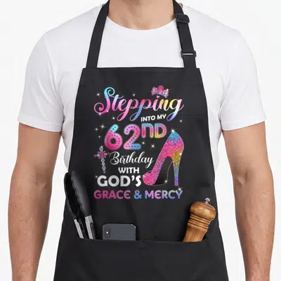 62nd birthday apron - Glittery high heel, rainbow text Stepping Into My 62nd Birthday With Gods Grace And Mercy, crown — shown on a black cotton canvas apron against a white background