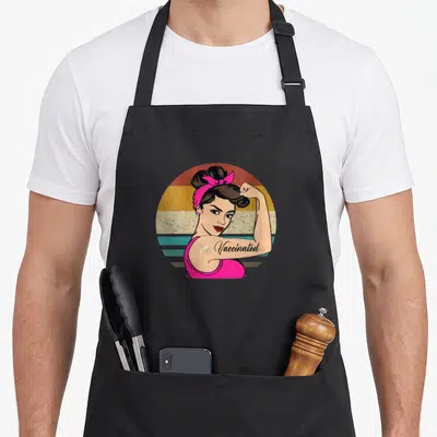 Vaccinated vaccine pro apron - Retro woman flexing arm with Vaccinated text and bandage on multi-colored striped circle — shown on a black cotton canvas apron against a white background
