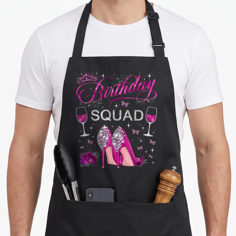 Heel butterfly apron - Birthday Squad text with high heels, wine glasses, butterflies and roses in pink and silver — shown on a black cotton canvas apron against a white background