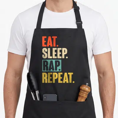 Eat rapper apron - Stacked EAT SLEEP RAP REPEAT text in red beige teal and yellow — shown on a black cotton canvas apron against a white background