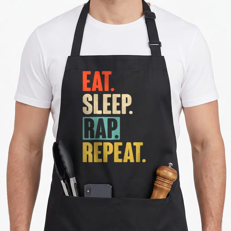 Eat rapper apron - Stacked EAT SLEEP RAP REPEAT text in red beige teal and yellow — shown on a black cotton canvas apron against a white background