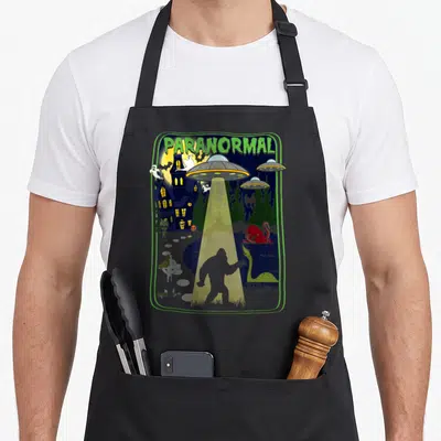 Paranormal bigfoot apron - Cartoon scene of Bigfoot under UFO beam near haunted house with ghosts and lake monsters — shown on a black cotton canvas apron against a white background