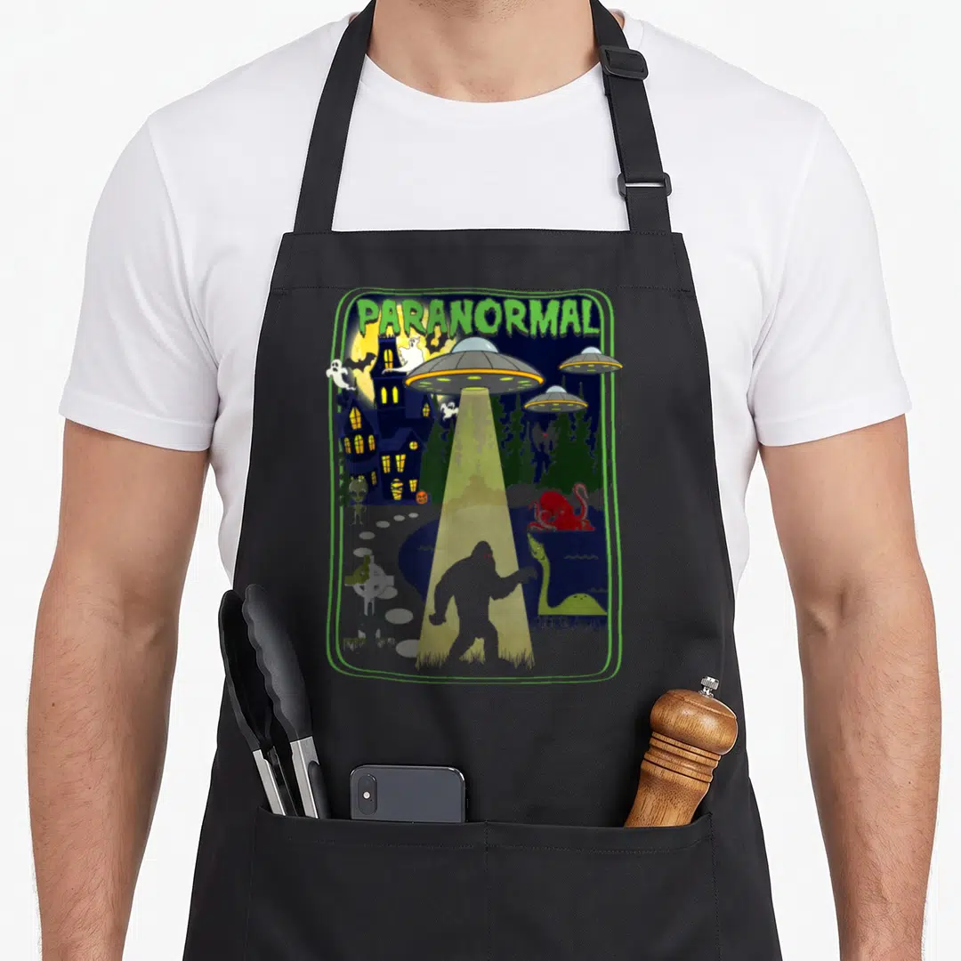 Paranormal bigfoot apron - Cartoon scene of Bigfoot under UFO beam near haunted house with ghosts and lake monsters — shown on a black cotton canvas apron against a white background