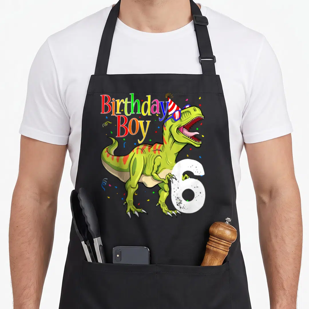 6th dinosaur apron - Cartoon T-Rex with party hat glasses number 6 and Birthday Boy text in rainbow colors — shown on a black cotton canvas apron against a white background