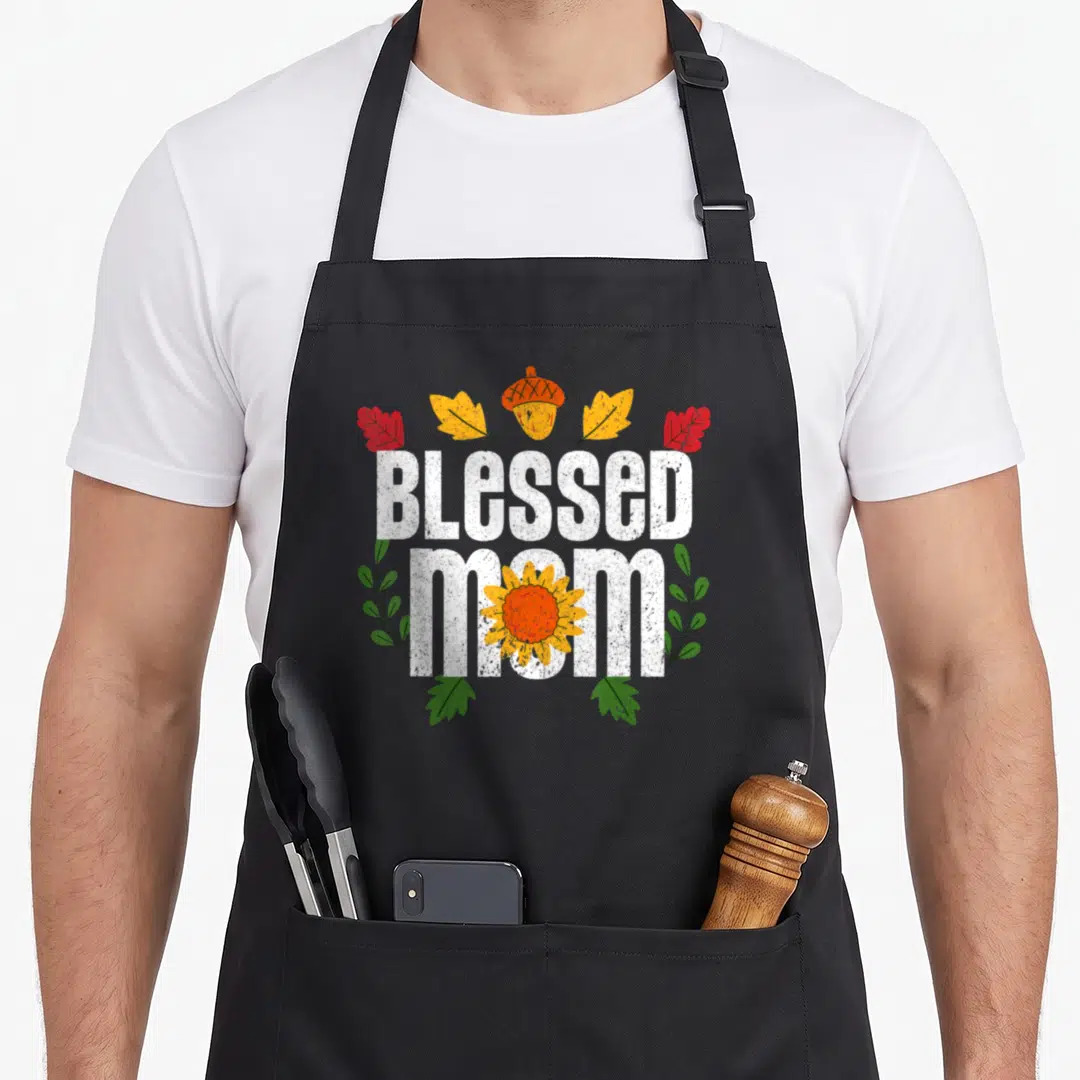Pilgrim thanksgiving apron - BLESSED MOM text with acorn, sunflower, four leaves, and green branches in distressed style — shown on a black cotton canvas apron against a white background