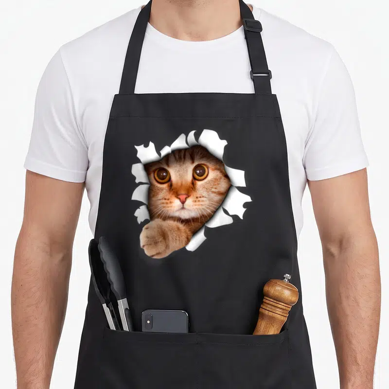 Realistic pet apron - Cat face and paw emerging through torn white paper in brown and orange photo style — shown on a black cotton canvas apron against a white background