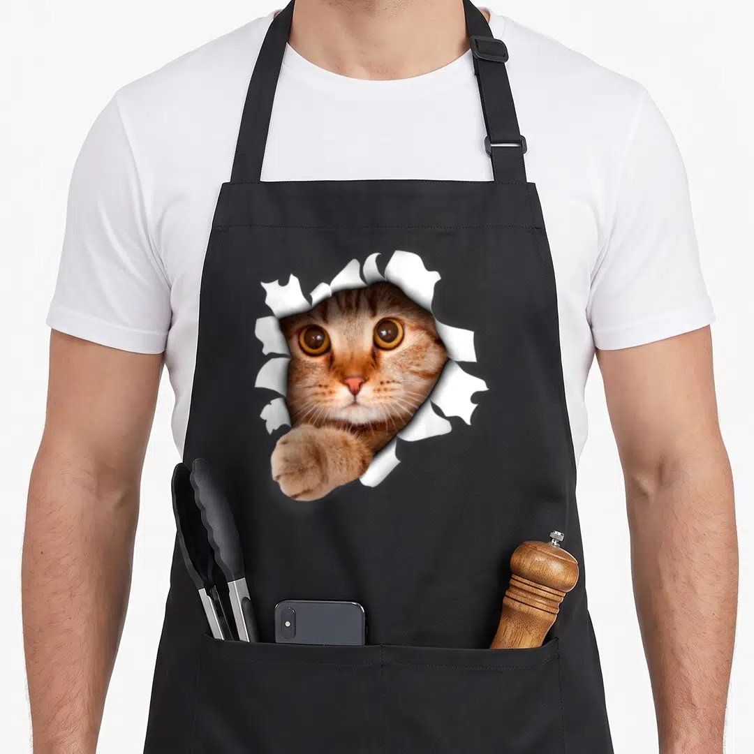 Realistic pet apron - Cat face and paw emerging through torn white paper in brown and orange photo style — shown on a black cotton canvas apron against a white background