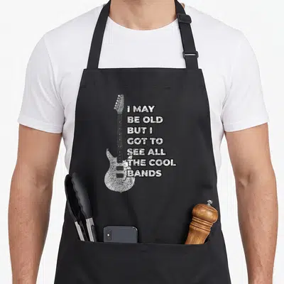 Vintage rock 80s apron - Distressed white electric guitar next to seven lines of stacked black text on a dark background — shown on a black cotton canvas apron against a white background