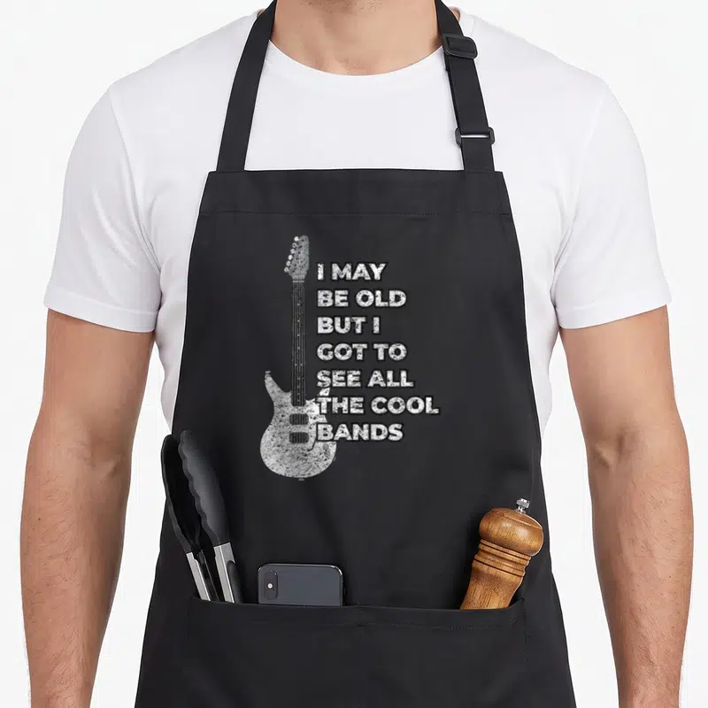 Vintage rock 80s apron - Distressed white electric guitar next to seven lines of stacked black text on a dark background — shown on a black cotton canvas apron against a white background