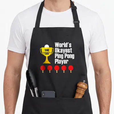 Okayest ping pong apron - Yellow trophy with YOU TRY text, five red paddles, and World's Okayest Ping Pong Player — shown on a black cotton canvas apron against a white background