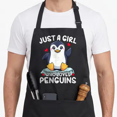 Funny animal penguin aprons - Cartoon penguin on ice floe with hearts and text Just a Girl Who Loves Penguins — shown on a black cotton canvas apron against a white background