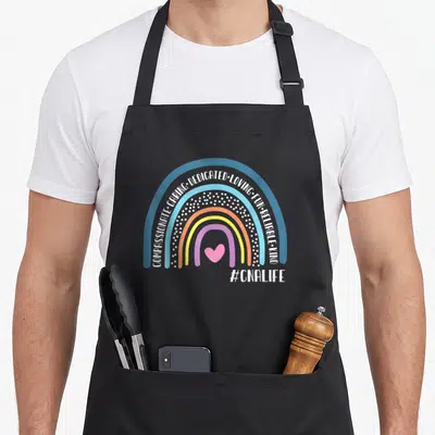 Assistant nursing aprons - Rainbow with blue orange purple pink bands containing white text and a pink heart below — shown on a black cotton canvas apron against a white background