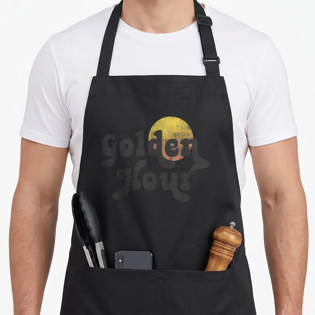 Trendy golden apron - Retro grey text Golden Hour with yellow sun graphic on black background — shown on a black cotton canvas apron against a white background