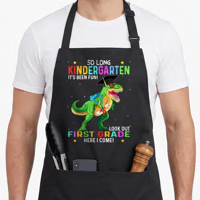 Kindergarten dinosaur apron - Green T-Rex in graduation cap holding diploma with colorful text celebrating kindergarten — shown on a black cotton canvas apron against a white background