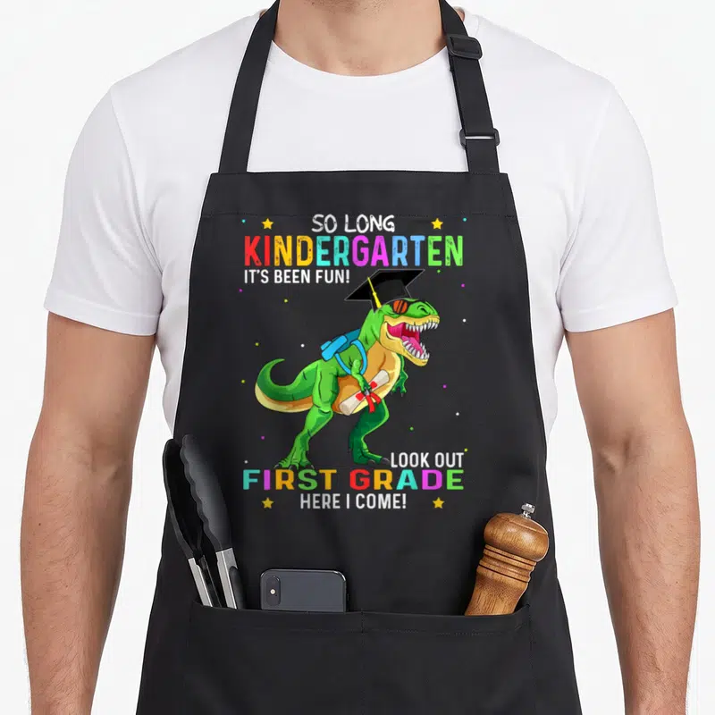 Kindergarten dinosaur apron - Green T-Rex in graduation cap holding diploma with colorful text celebrating kindergarten — shown on a black cotton canvas apron against a white background