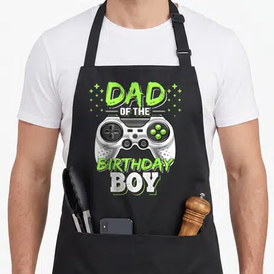 Video gamer birthday apron - Dad of the Birthday Boy text above white game controller with lime green plus signs — shown on a black cotton canvas apron against a white background