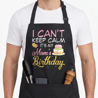 Balloon cake apron - I Can't Keep Calm It's My Mom Birthday text with cake and balloons in pink gold white — shown on a black cotton canvas apron against a white background