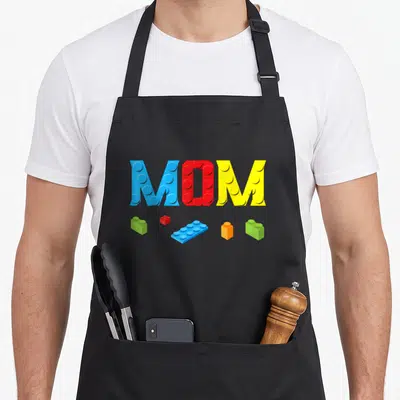 Builder block & apron - MOM built from colored building blocks with five scattered bricks below in cartoon style — shown on a black cotton canvas apron against a white background