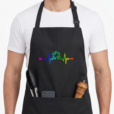 Beat teacher art apron - Rainbow heartbeat line with central puzzle piece and hearts in blue green yellow red pink — shown on a black cotton canvas apron against a white background
