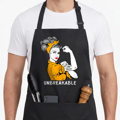 Warrior manuscript apron - Cartoon woman flexing arm with Ms Warrior tattoo and UNBREAKABLE text in yellow, white — shown on a black cotton canvas apron against a white background