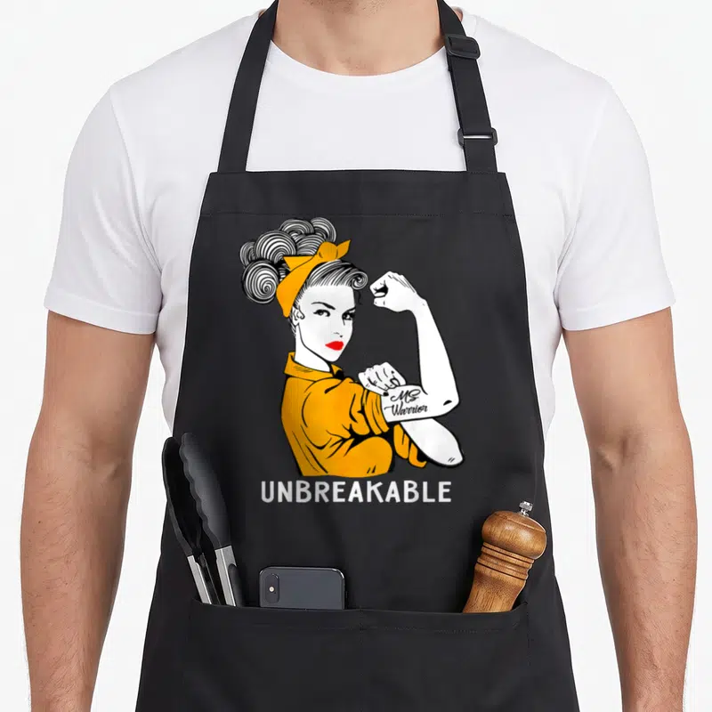 Warrior manuscript apron - Cartoon woman flexing arm with Ms Warrior tattoo and UNBREAKABLE text in yellow, white — shown on a black cotton canvas apron against a white background