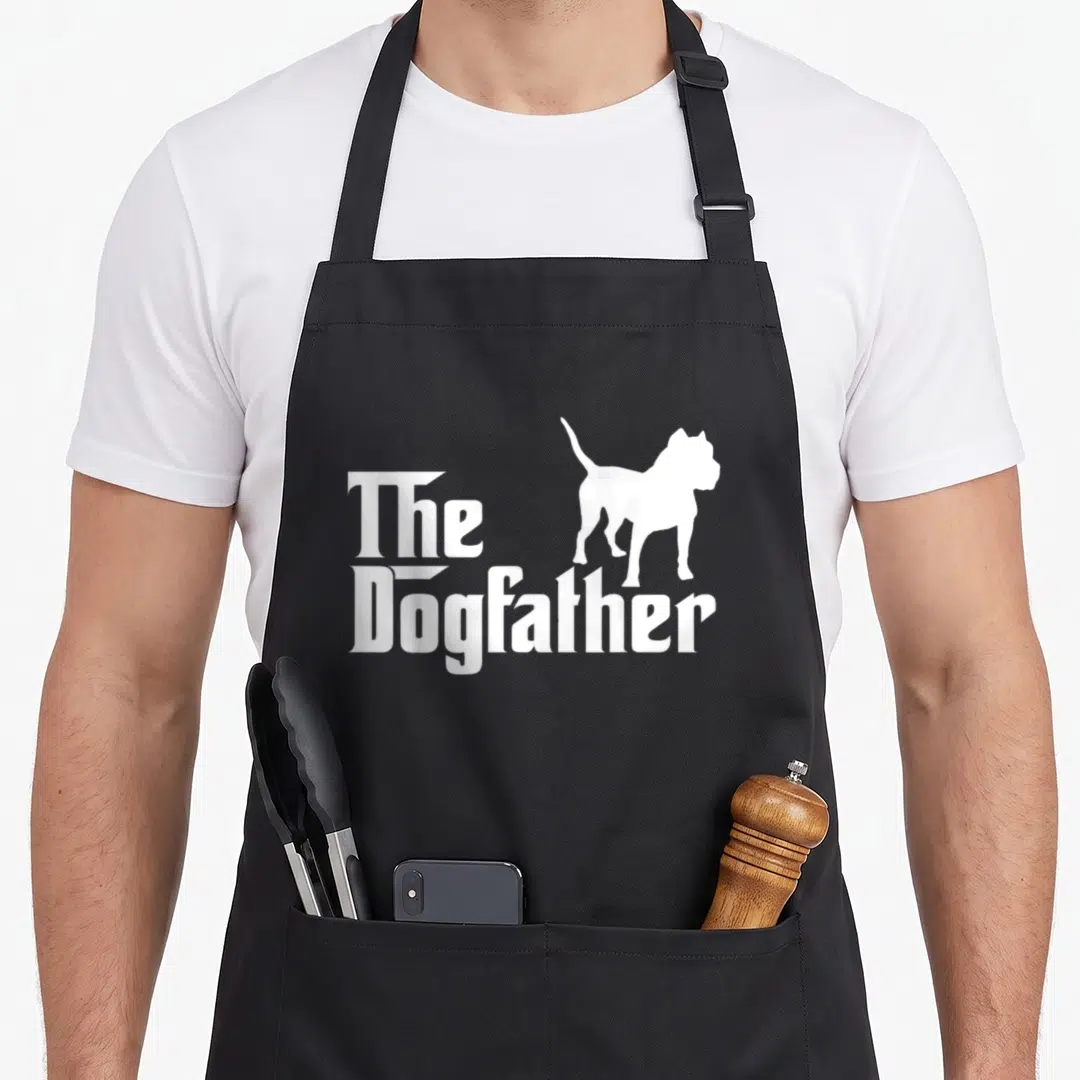Dogfather class funny apron - White stacked text reading The Dogfather next to a standing dog silhouette — shown on a black cotton canvas apron against a white background