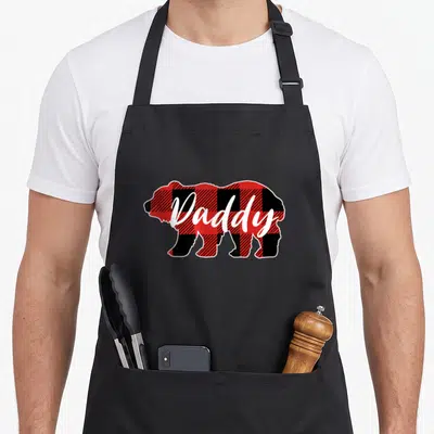 Daddy buffalo apron - Bear silhouette filled with red black and white plaid pattern with white Daddy script text overlay — shown on a black cotton canvas apron against a white background
